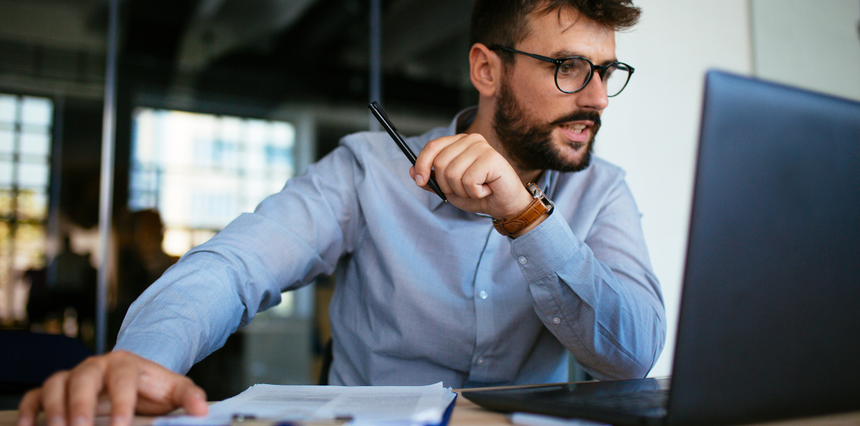 A dark haired man with a beard is wearing glasses and a blue shirt. He is working on a silver laptop.